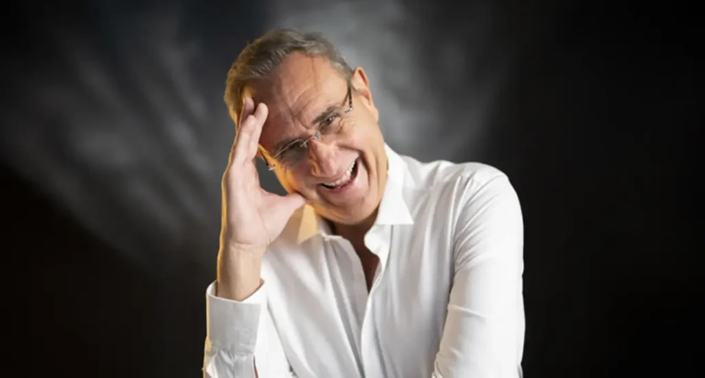 Uomo sorridente in camicia bianca, ritratto su sfondo scuro, con una mano appoggiata alla fronte in un gesto informale e ironico.