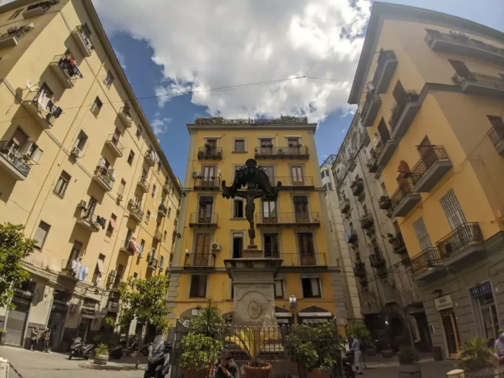 Piazza del Borgo Orefici a Napoli con la statua centrale, circondata da palazzi storici e botteghe artigiane.