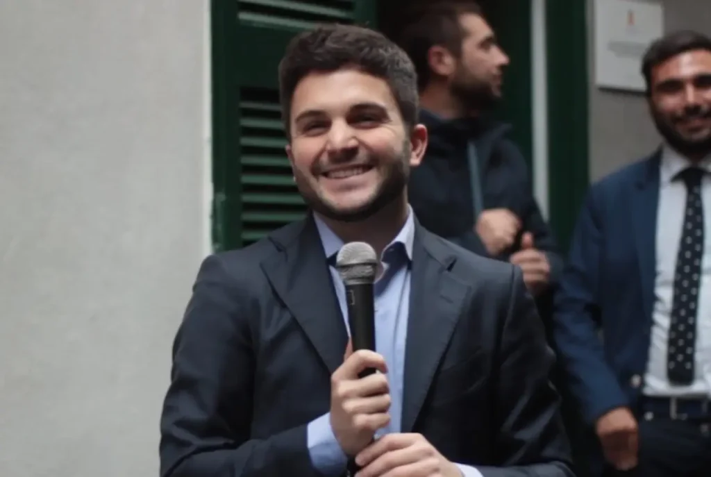Uomo in giacca scura tiene un microfono e sorride durante un intervento pubblico, con altre persone sullo sfondo.